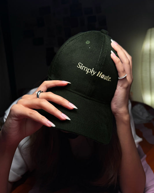 Person holding a olive green corduroy cap with 'Simply Haute' text against a dark background