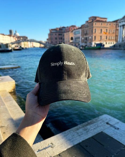 Olive green corduroy cap with 'Simply Haute' embroidered text held over a canal with buildings and a bridge in the background of Venice